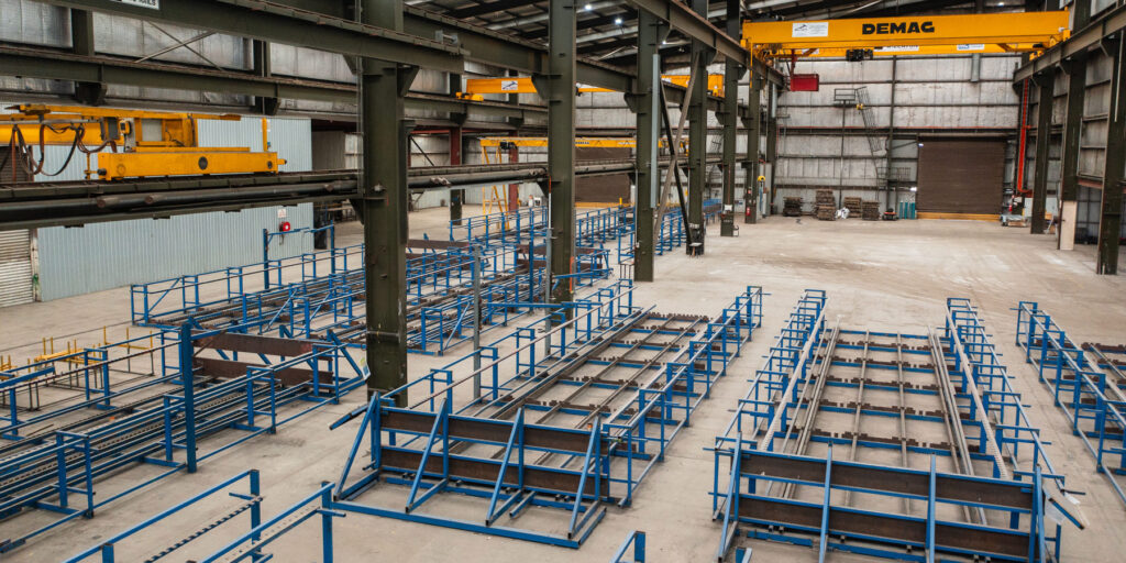 Inside of a very large industrial shed with numerous heavy duty overhead cranes, and blue racks storing long steel reinforcing rods.