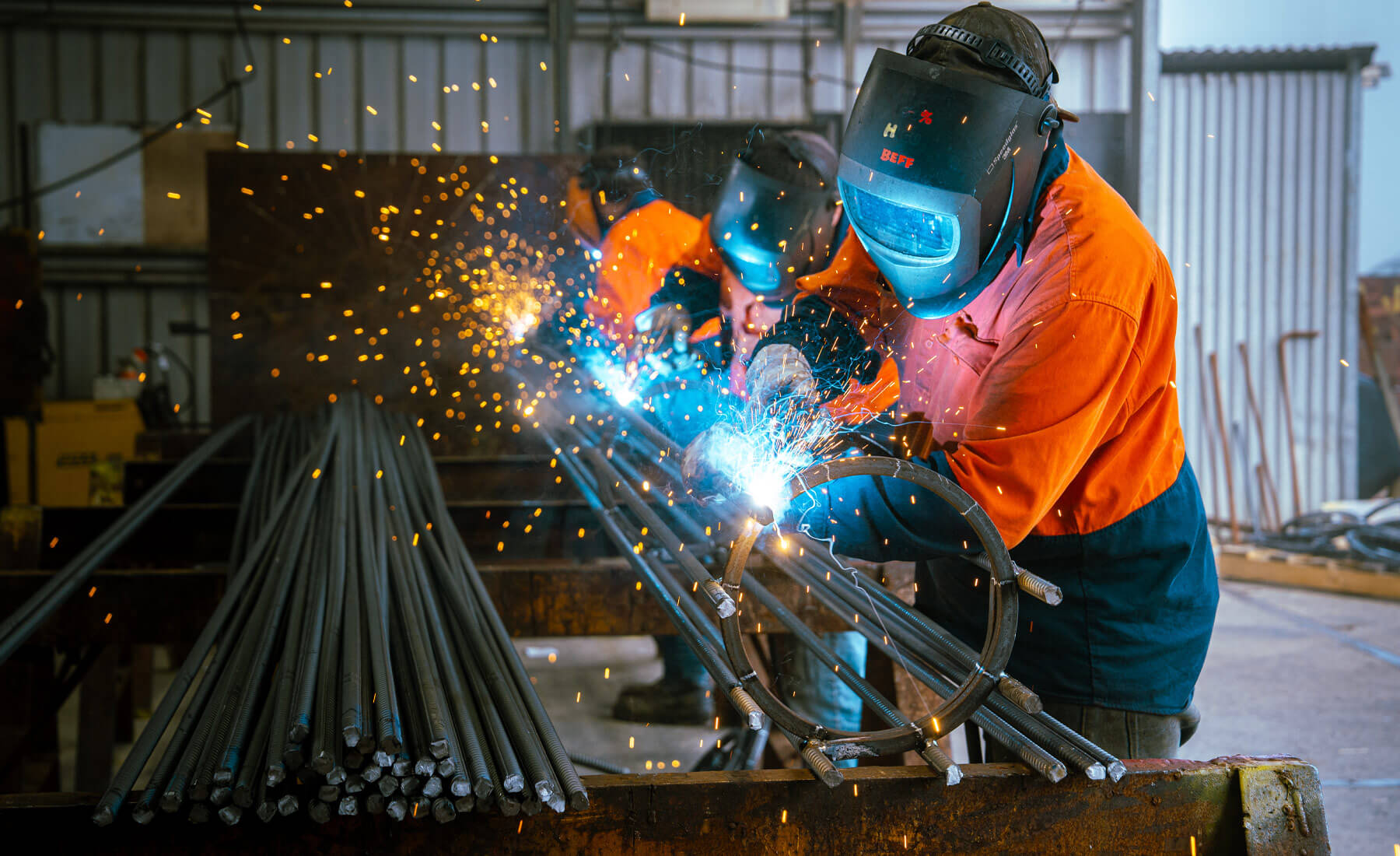 Three men working in a shed, welding steel reinforcing into a mesh column. The men are wearing welding masks, orange high-vis shirts, and the welding sparks are bright and creating interesting sparks and patterns of bright light.