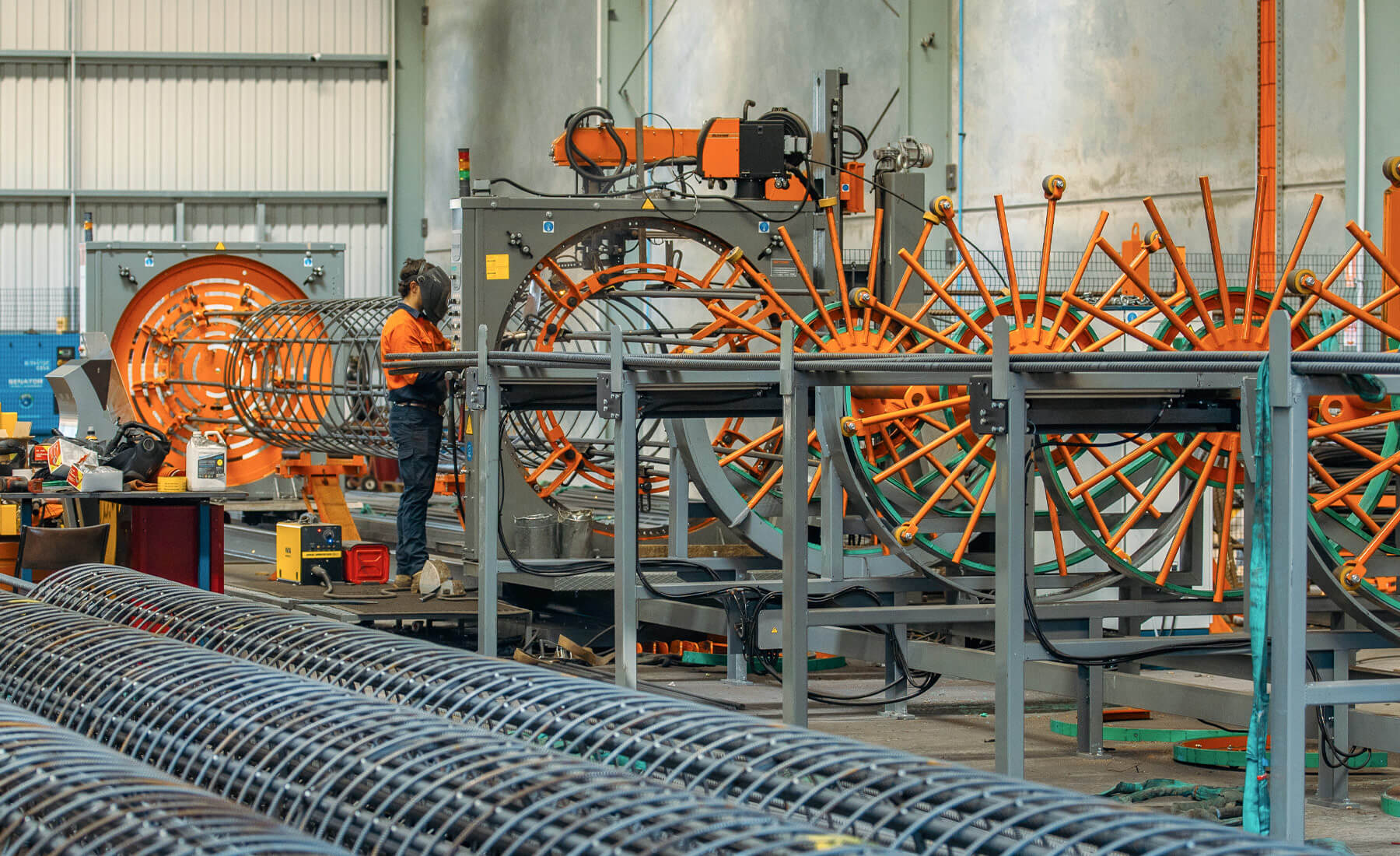 Photo of man in orange high-vis shirt wearing a welding mask and welding a steel circular reinforcement cage. A very complex machine is helping bend the steel rods into the circular shape.