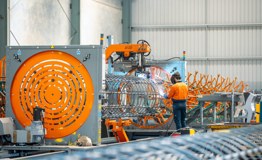 Photo of man in orange high-vis shirt wearing a welding mask and welding a steel circular reinforcement cage.