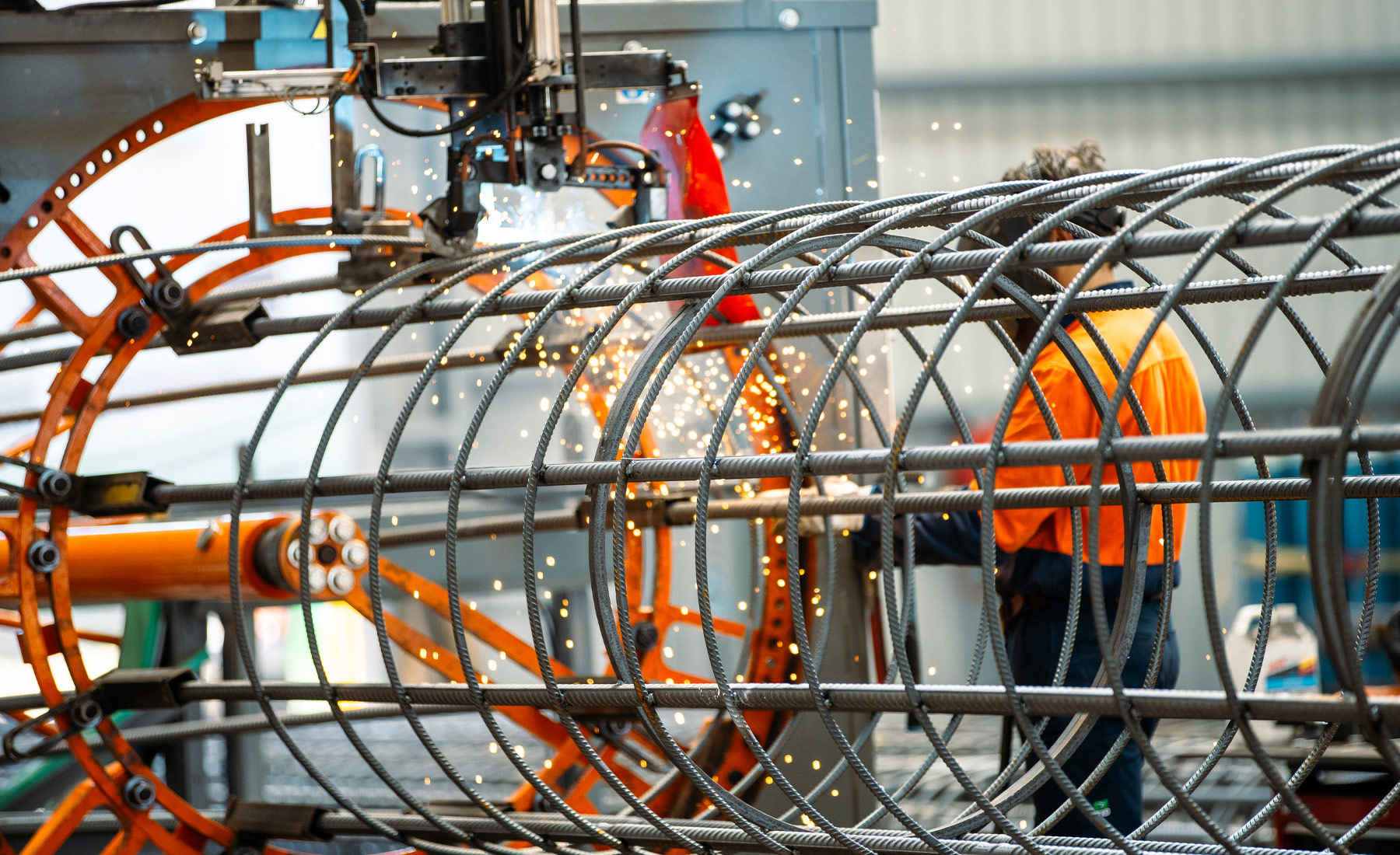 Photo of man in orange high-vis shirt wearing a welding mask and welding a steel circular reinforcement cage.