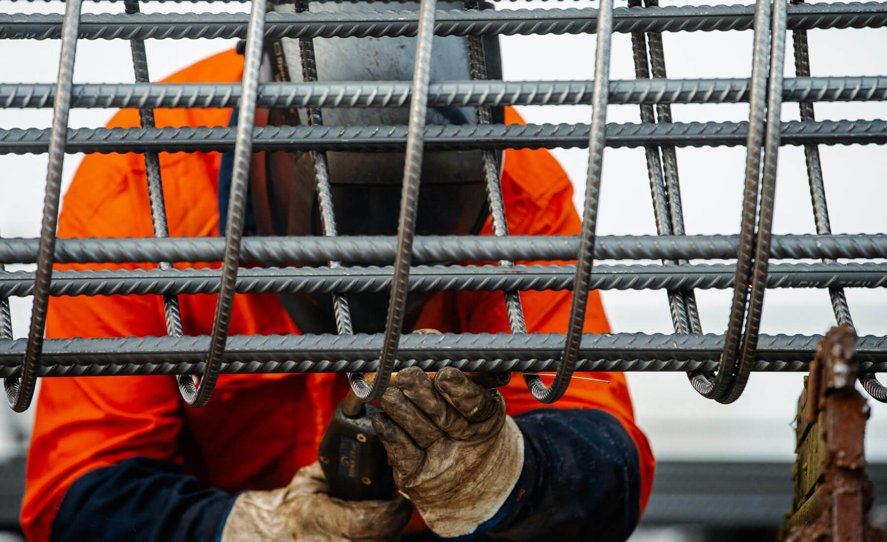 Man with welding mask on and wearing an orange high-vis shirt and thick gloves as he works on a mesh column made from steel rod reinforcing.
