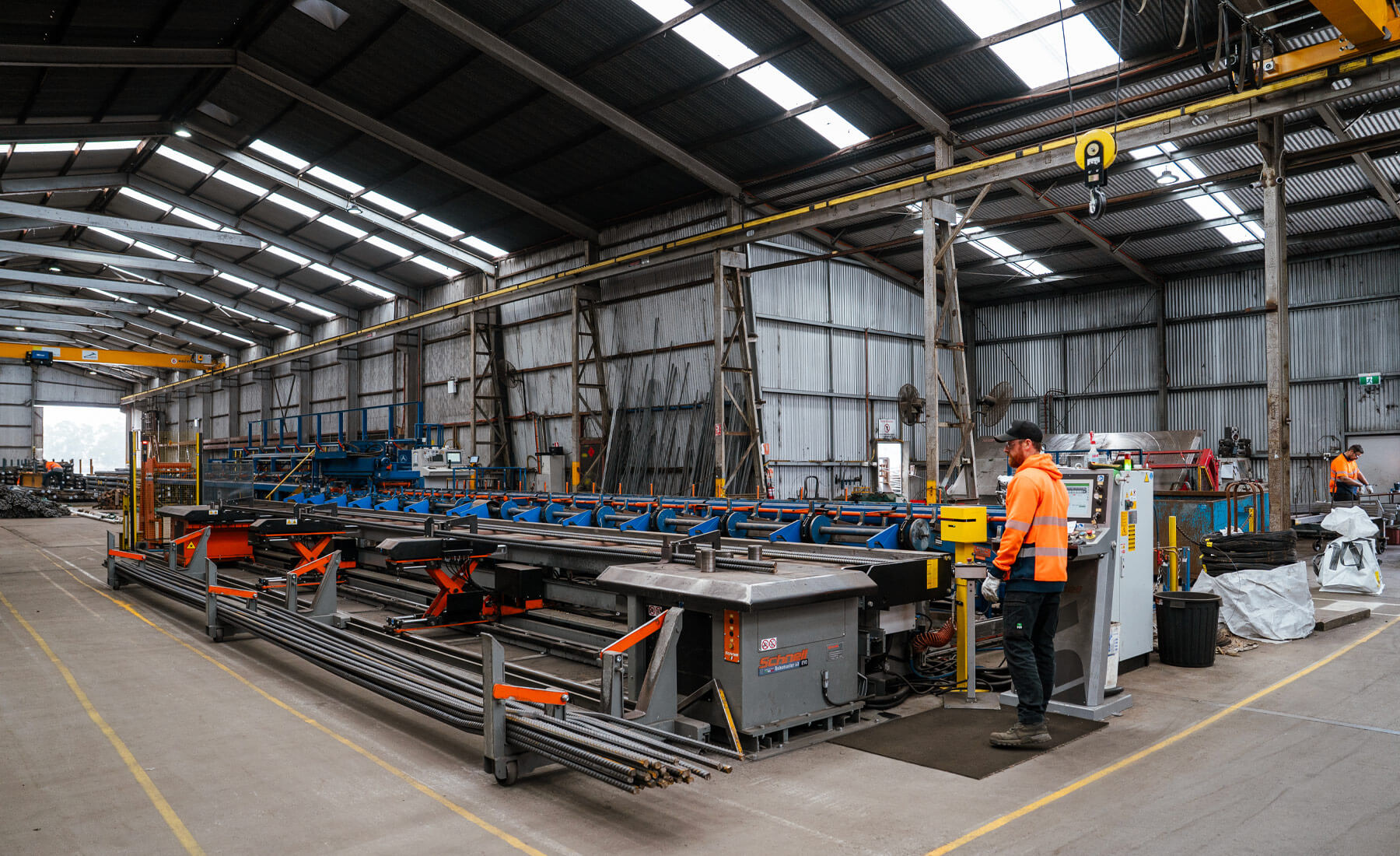 Man with a beard, wearing a brown cap, safety glasses and high-vis orange jacket, orperating a large machine in a big shed. The machine is used to bend steel reinforcing bars and is a very long machine.