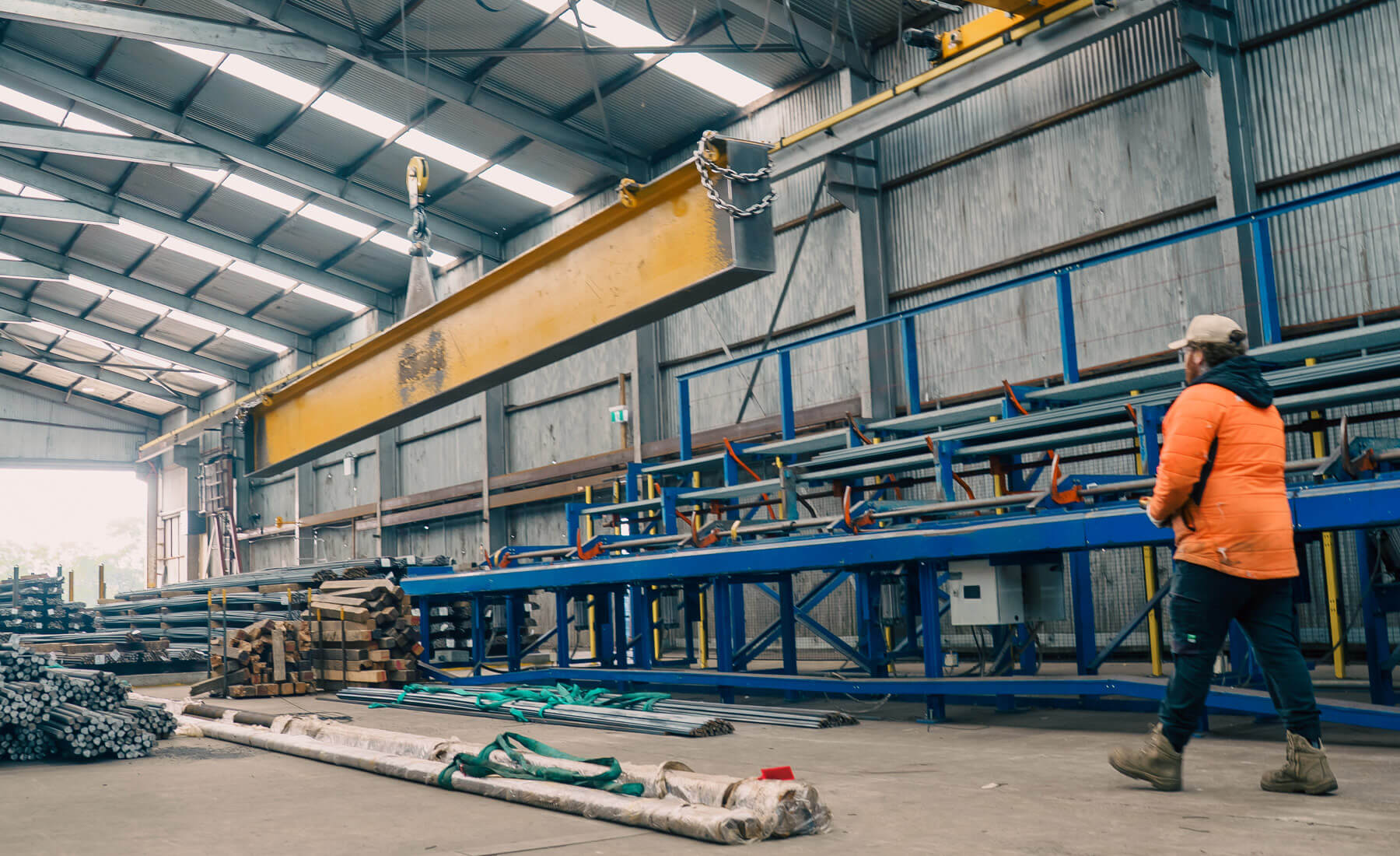 Man in a workshop wearing a high-vis nacket as he uses a large overhead crane to life a large steel beam. The crane has a large chain and hook and is operated by remote.