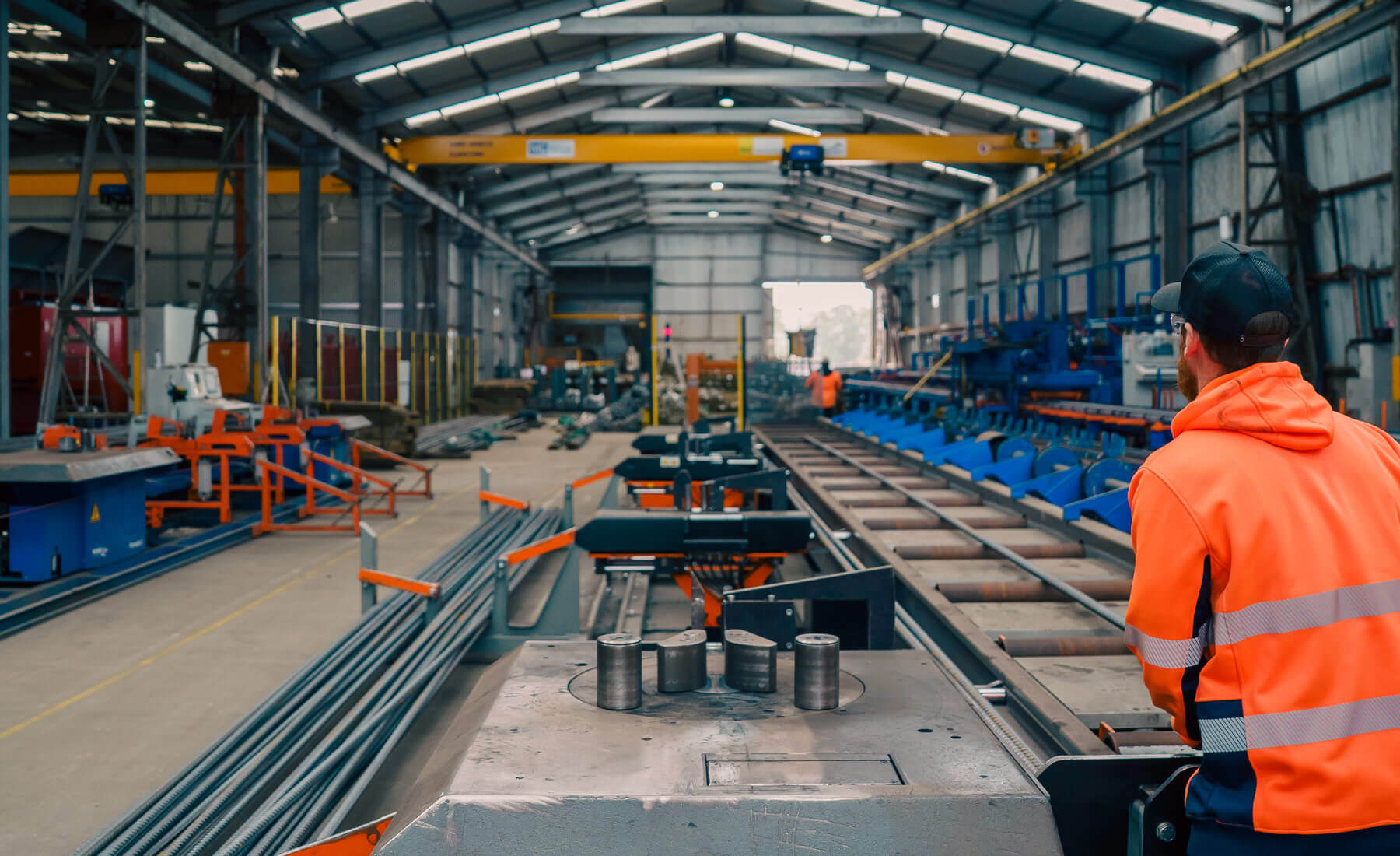 Looking over the should of a man wearing a cap and high-vis shirt as he operates a long machine that bends steel reinforcing bars.