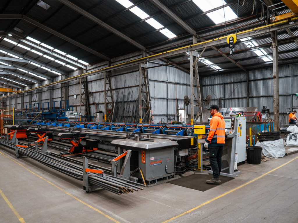 Man with a beard, wearing a brown cap, safety glasses and high-vis orange jacket, orperating a large machine in a big shed. The machine is used to bend steel reinforcing bars and is a very long machine.