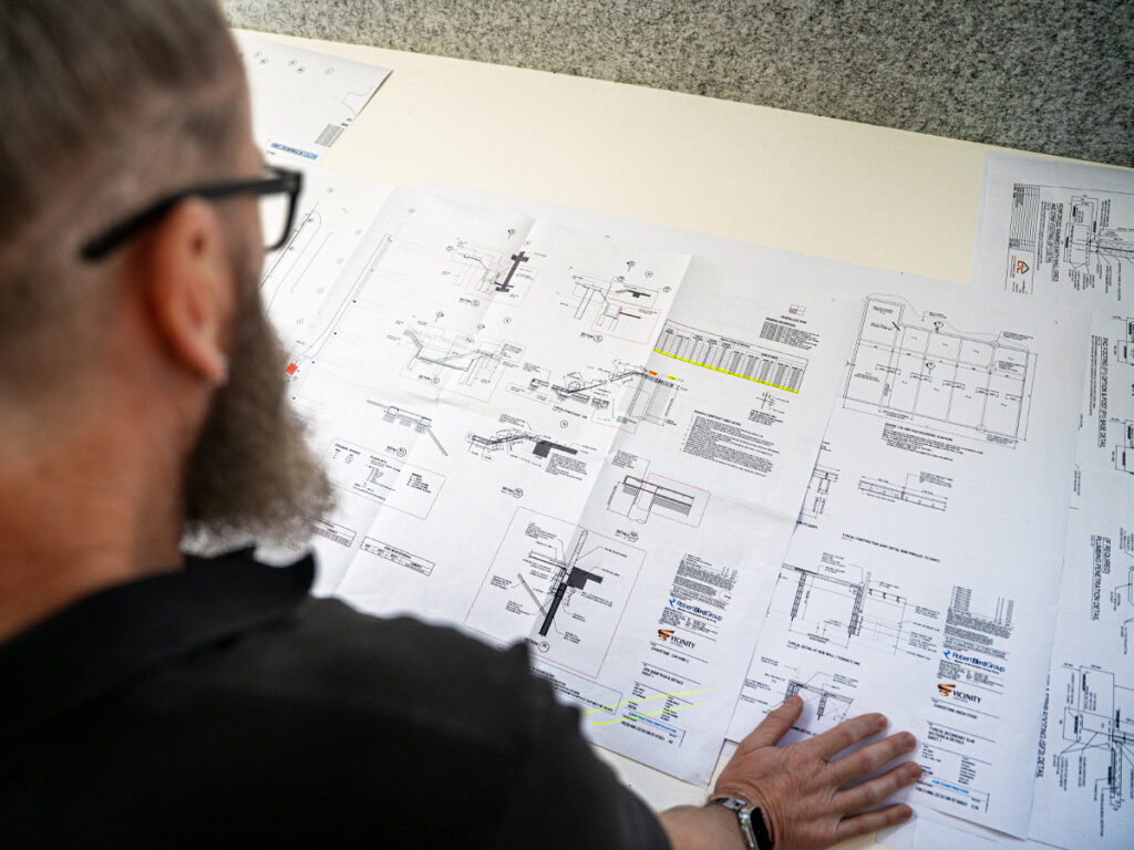 Looking over the shoulder of a man as he inspects detailed planning drawings. The man has a beard and is wearing glasses and a black shirt.
