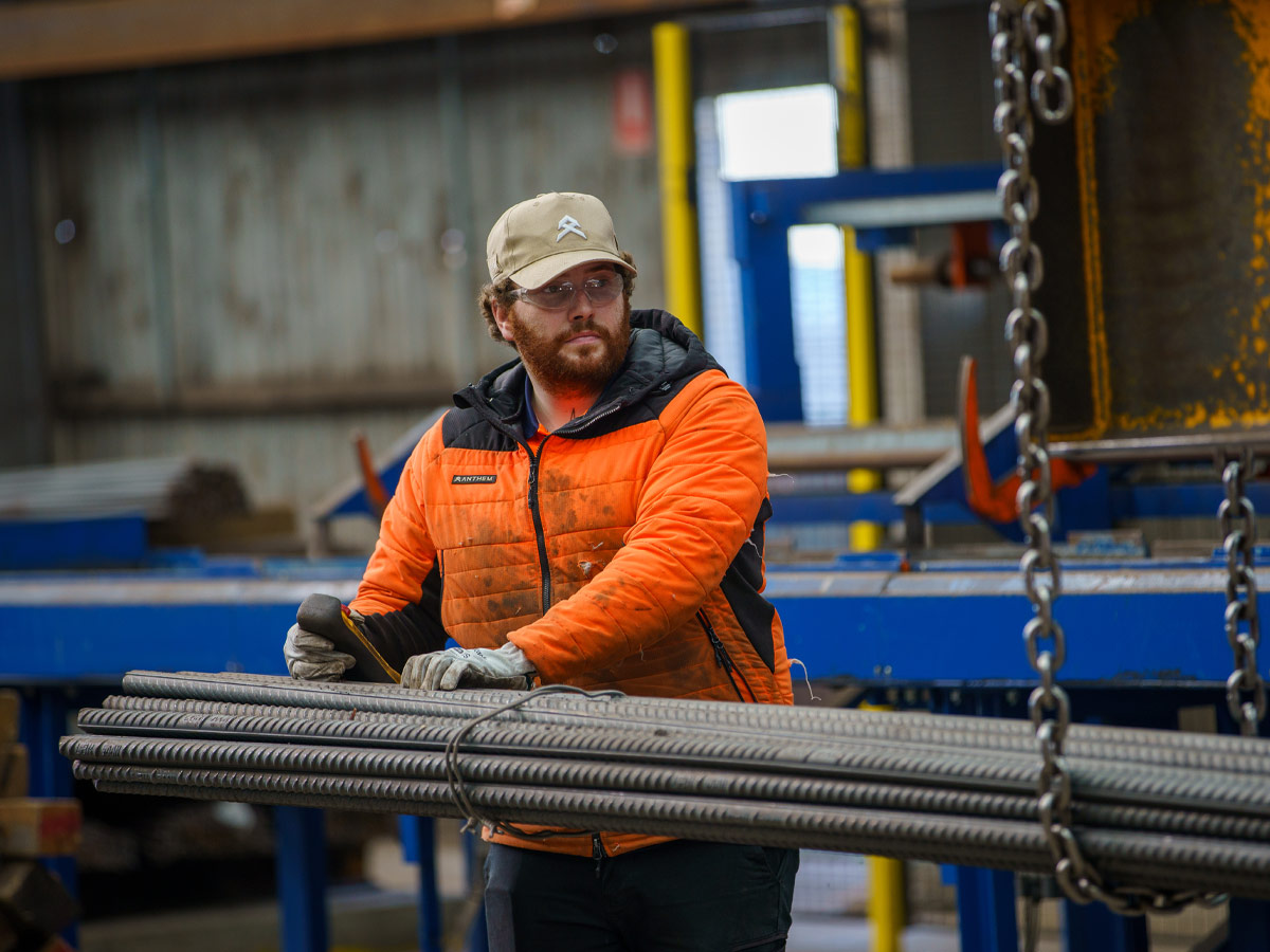 Man with a beard, wearing a brown cap, safety glasses and high-vis orange jacket, moving bars of steel reinforcing via a crane with chains.